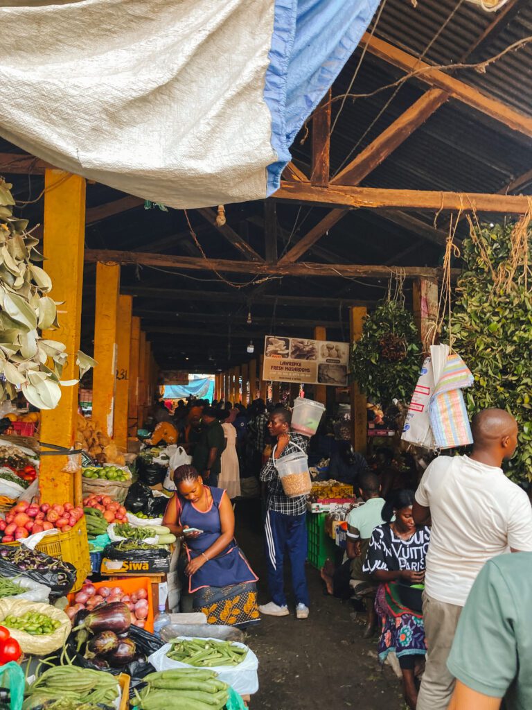 Een vrouw zit naast haar groentekraam op Nakaseero Market in Kampala, omringd door een bonte verzameling van verse groenten en fruit. Haar aandacht is gefocust op de kraam terwijl ze wacht op klanten, en haar gezicht straalt een gevoel van trots en toewijding uit.
