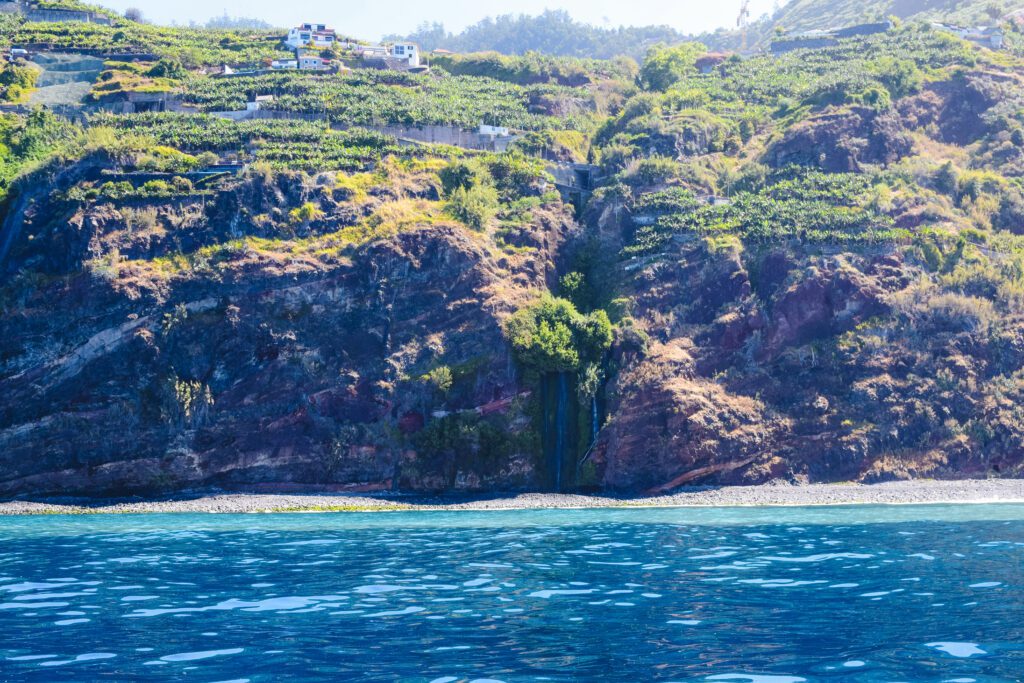 ingezoomde foto genomen van op zee richting het eiland. Er is een waterval zichtbaar op het verlaten stukje strand, dat verscholen zit in de rotswand.