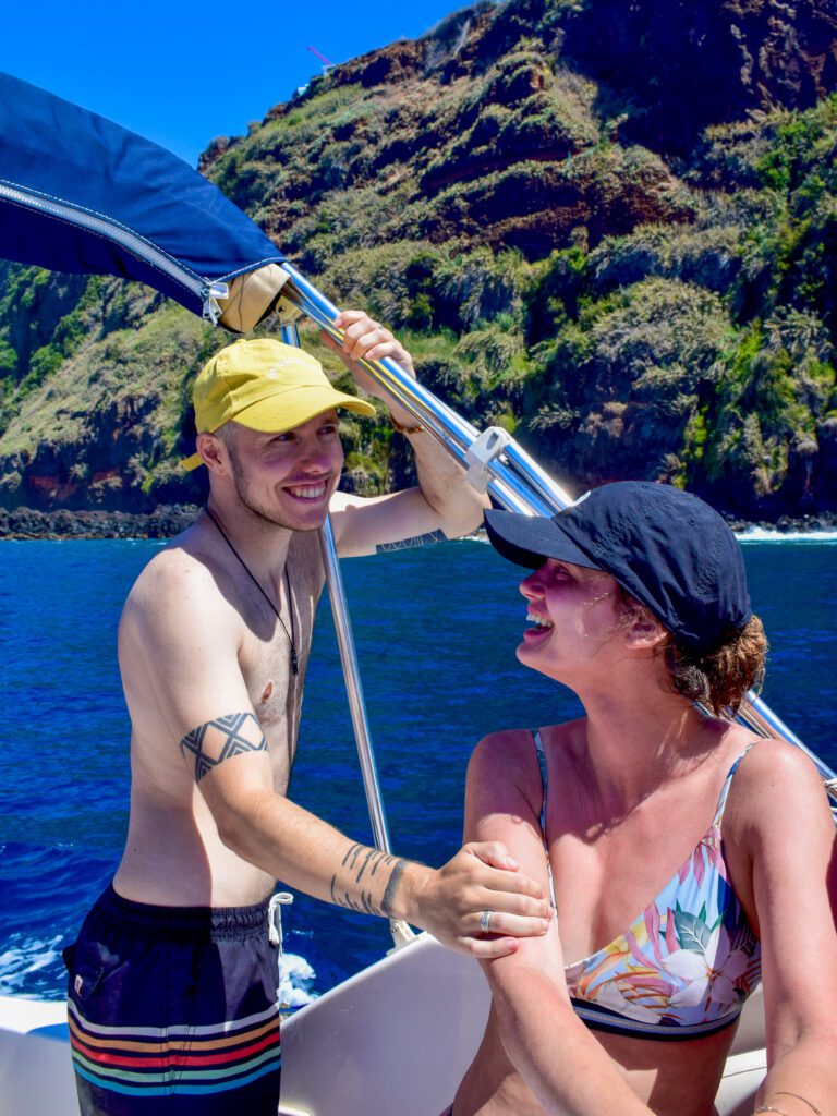 Davo staat links, draagt een zwemshort en een gele pet en houdt het dak van de boot vast met zijn ene hand, met zijn andere hand houdt hij de bovenarm van Lotte vast die een bikini draagt en een zwarte pet en naar hem lacht. Op de achtergrond zijn de kliffen van het eiland zichtbaar.