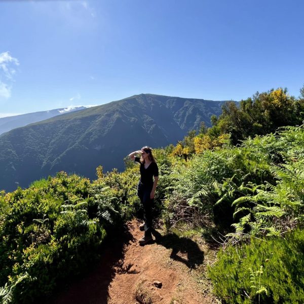 Lotte kijkt in de verte vanop miradouro Fio in Fanal Forest. Ze staat op enorm hoogte en kijkt uit over de kliffen, die bedenkt zijn met groene vegetatie en in de verte is de oceaan te zien.